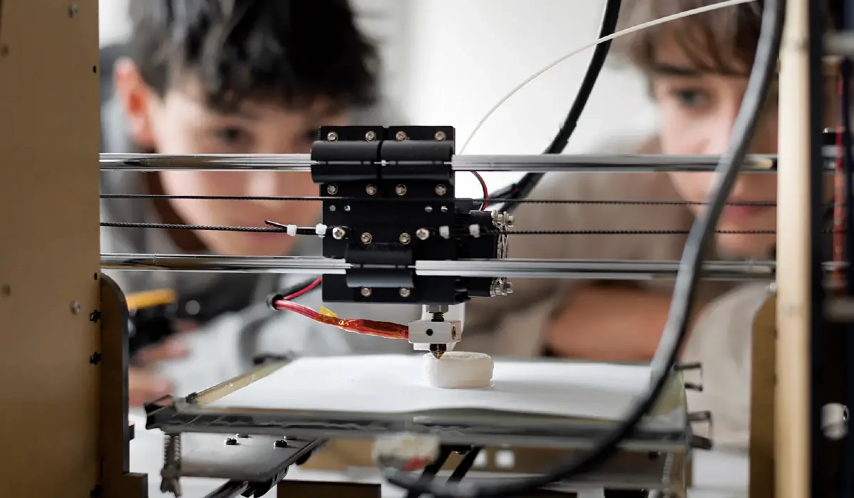 Two children watch intently as a 3D printer creates a round object from white filament. The scene demonstrates technological interest and curiosity in a learning environment.