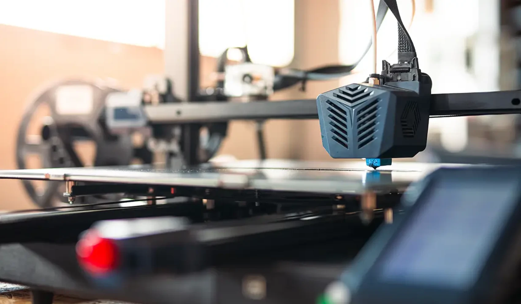 Ein moderner 3D-Drucker arbeitet präzise an einem Objekt, der Druckkopf ist aktiv in Bewegung. Im Hintergrund sind Filamentrollen und unscharfe technische Komponenten zu erkennen.
