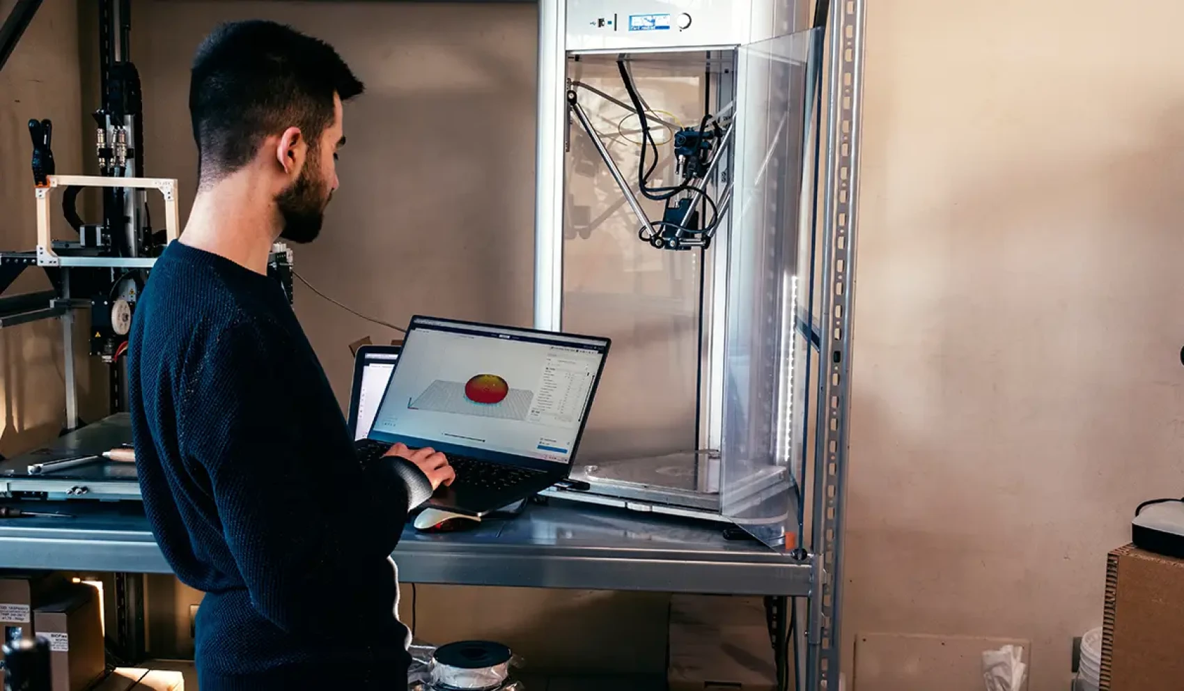 A man works on a laptop displaying a 3D model, while a large 3D printer sits next to him. The setting resembles a workshop or laboratory with technical equipment.